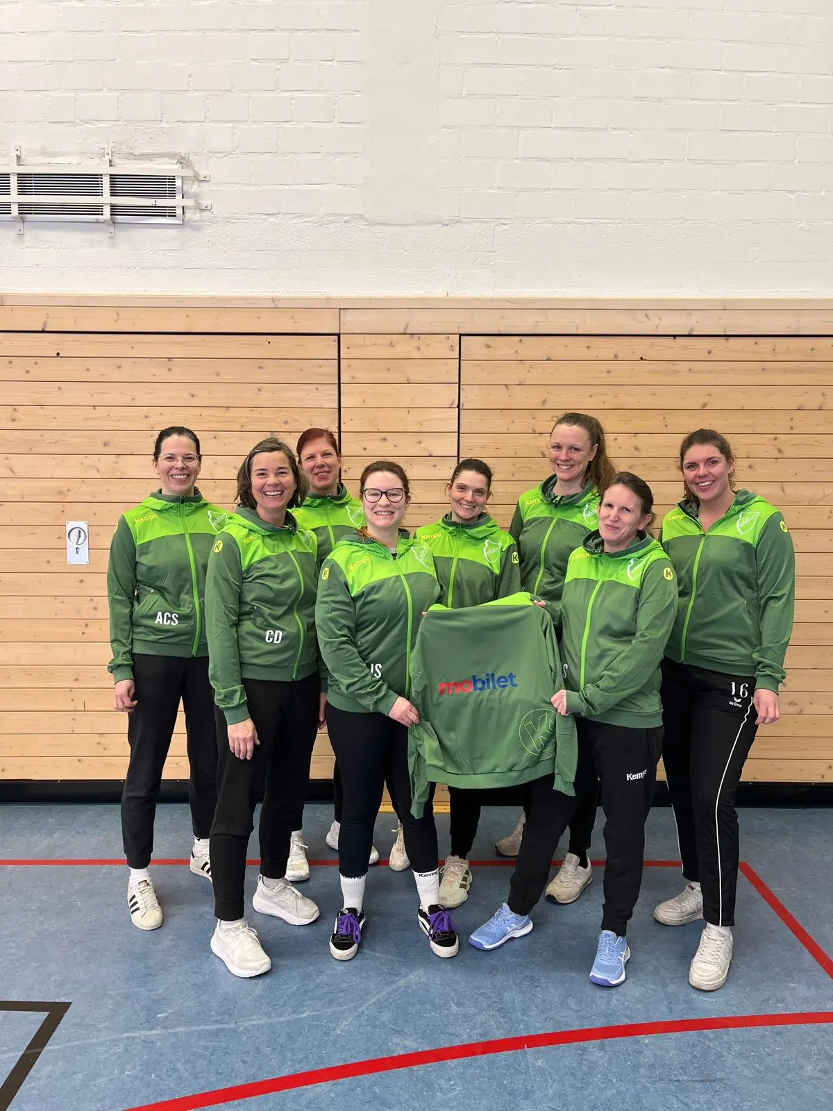 Gruppe von acht Frauen in grünen Trainingsjacken, die in einer Sporthalle stehen und in die Kamera lächeln.